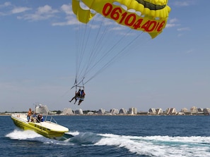 Ein Parasailing-Schirm in Gelb mit roter Schrift, ein Schnellboot mit der Aufschrift 'MACHUTE' und einer Telefonnummer, sowie eine Küstenstadt im Hintergrund.