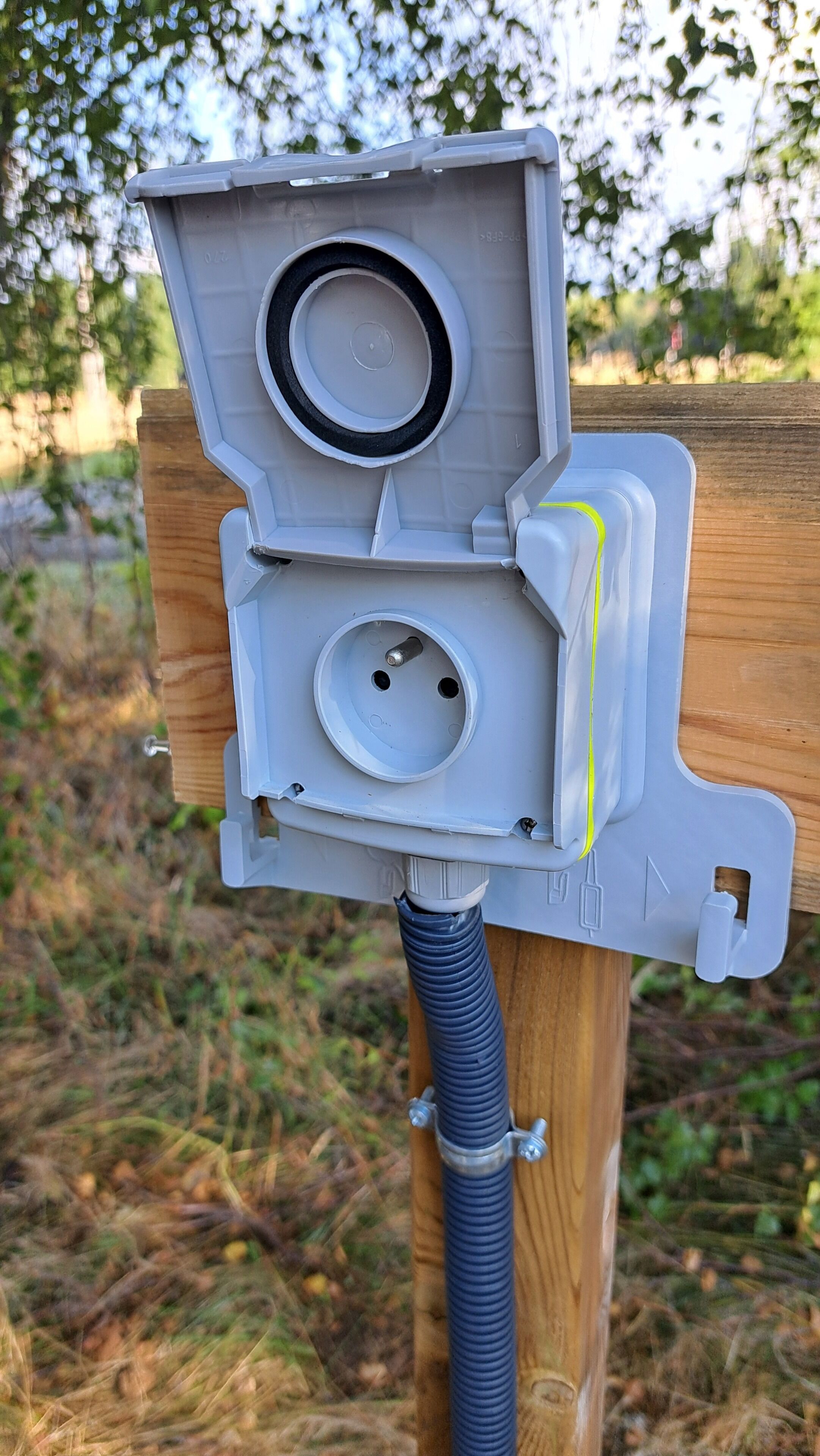 A utility box with a circular opening and a smaller circular opening below it, mounted on a wooden post with a blue hose attached.