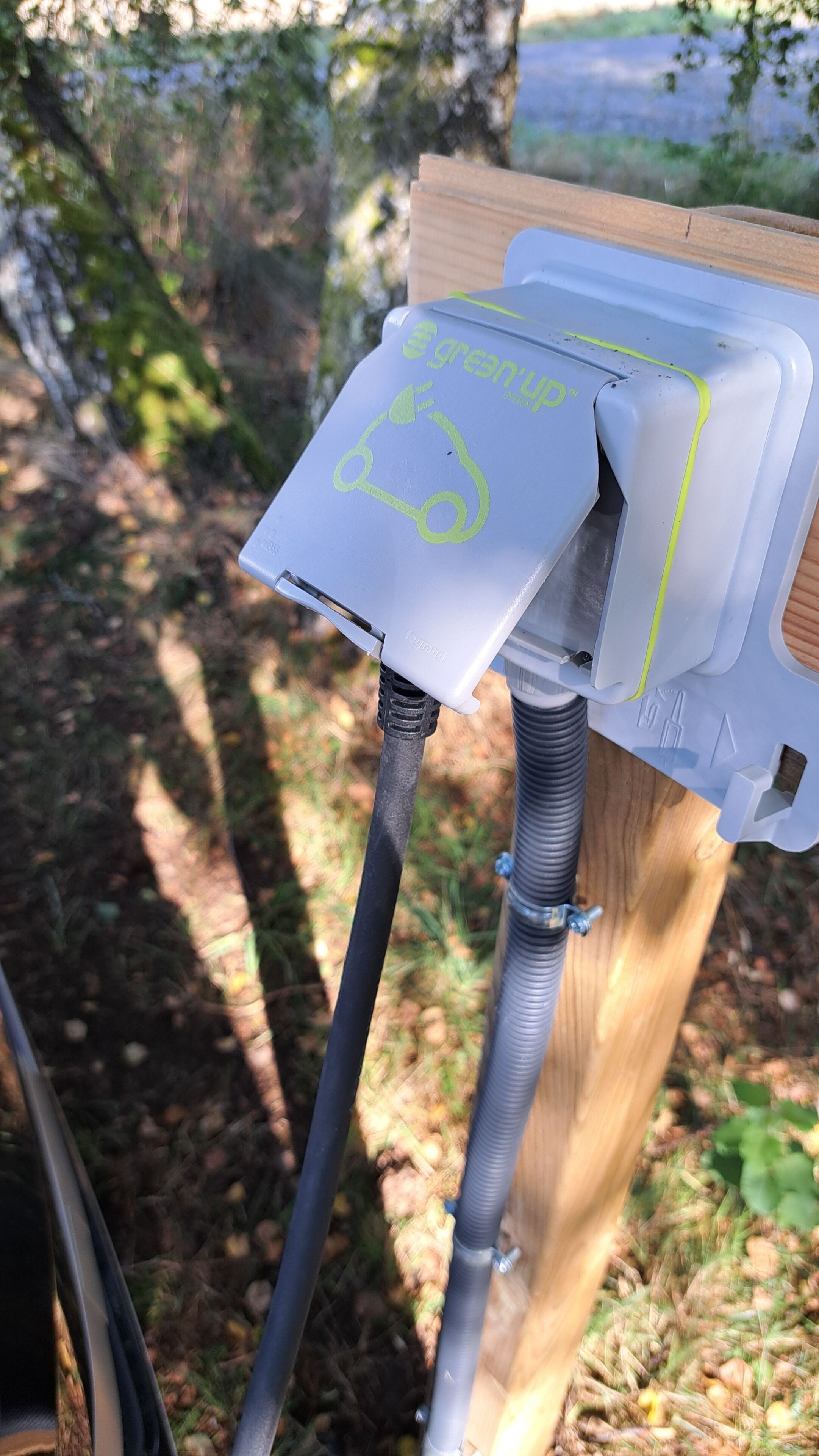 A close-up of a white electrical box with a green logo, attached to a wooden post with a black cable connected to it.