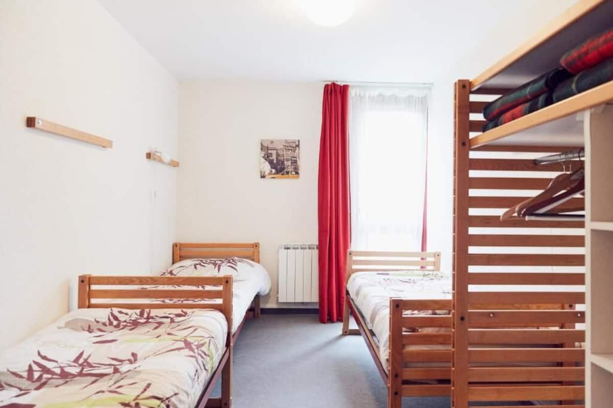 A dormitory room with two single beds, a radiator, a window with red curtains, and a wooden shelf with clothes.