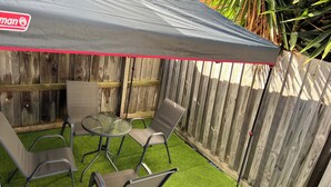 Een terras buiten met een grijze luifel, twee metalen stoelen, een kleine ronde tafel en een houten hek op de achtergrond.