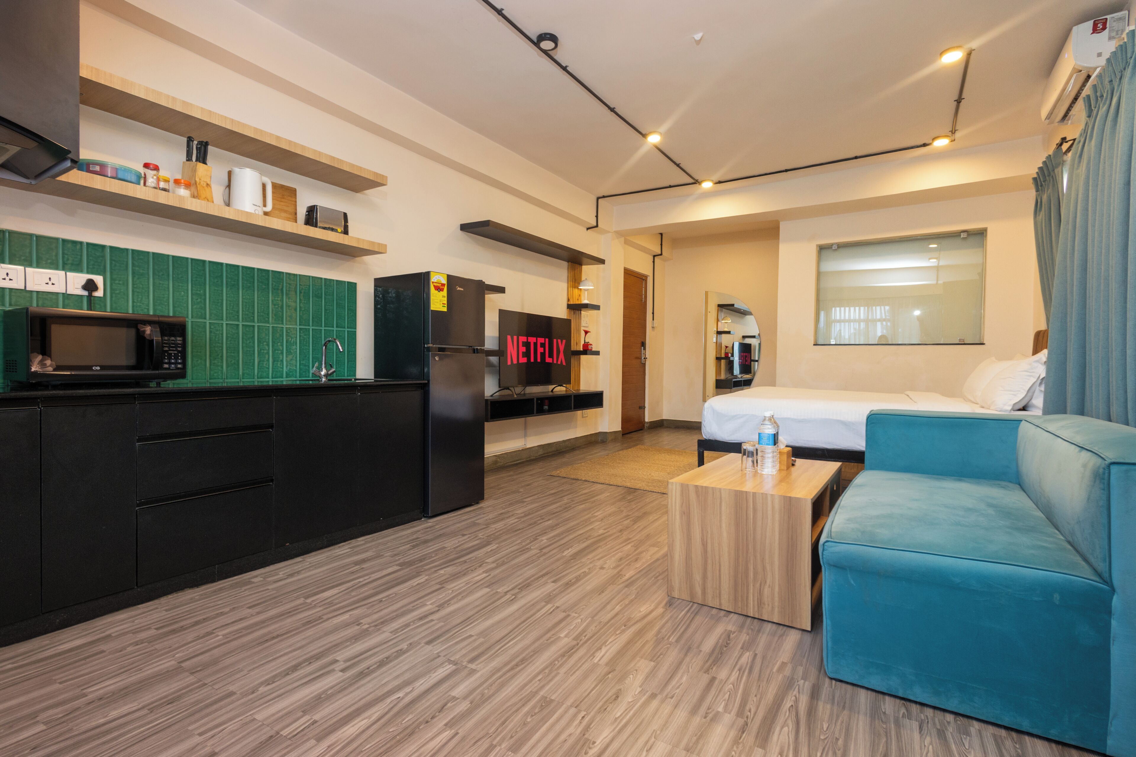 A modern hotel room with a kitchenette, a bed, a blue sofa, and a TV labeled 'NETFLIX'.