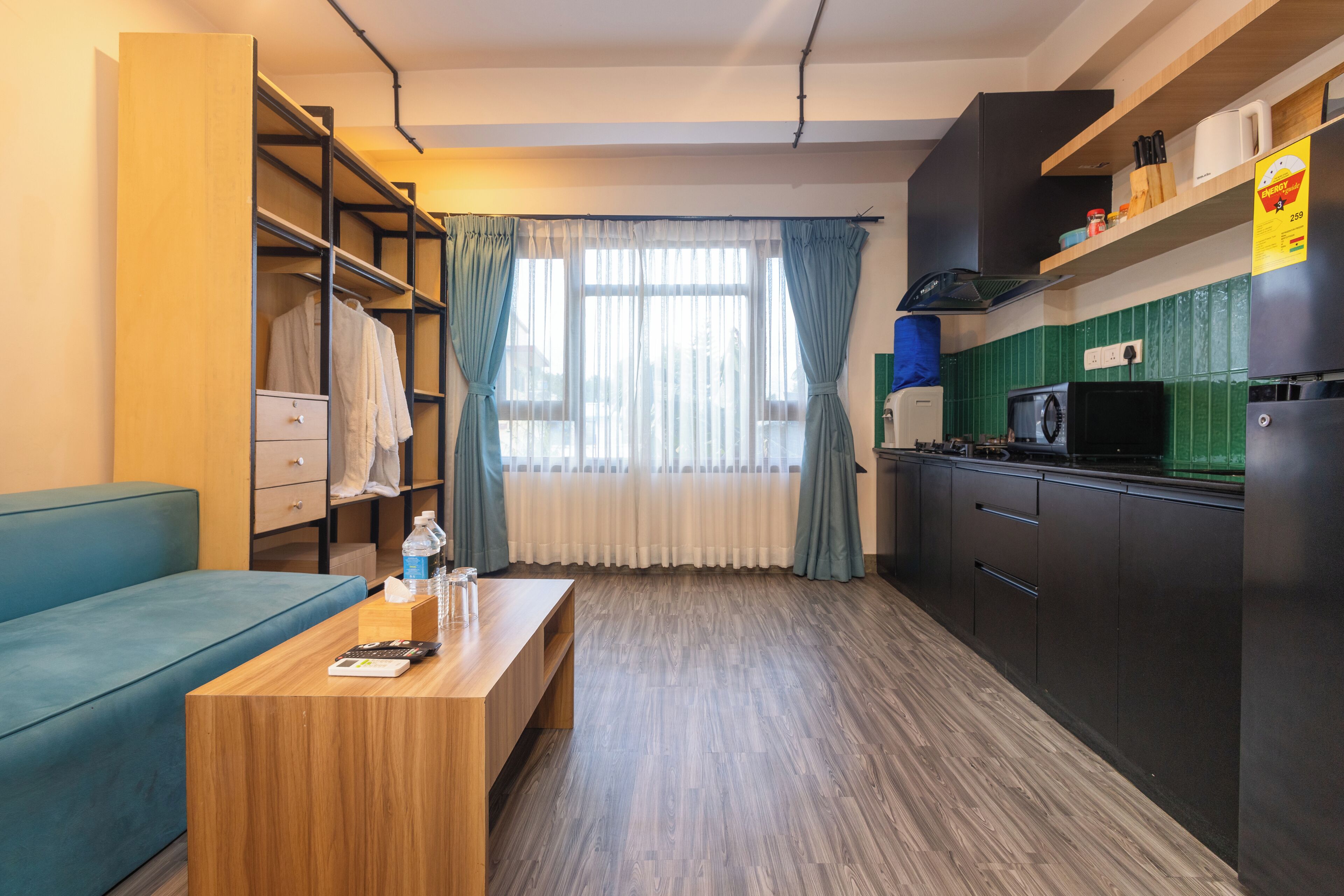 A compact hotel room with a kitchenette, a sofa, a wooden table with a remote control, and a window with curtains.