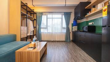 A compact hotel room with a kitchenette, a sofa, a wooden table with a remote control, and a window with curtains.