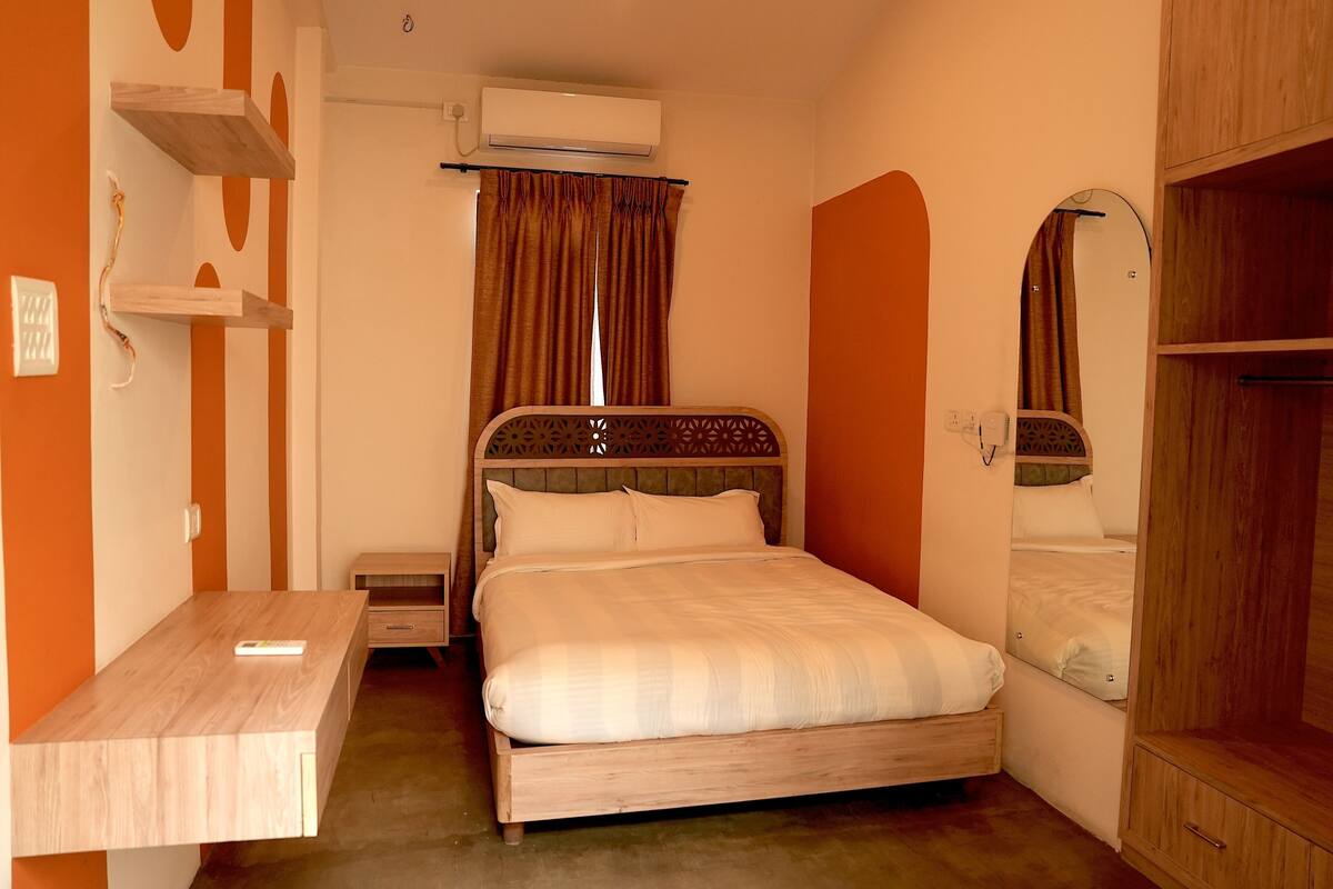 A bedroom with a bed, a wooden desk, a wardrobe, and an air conditioning unit.
