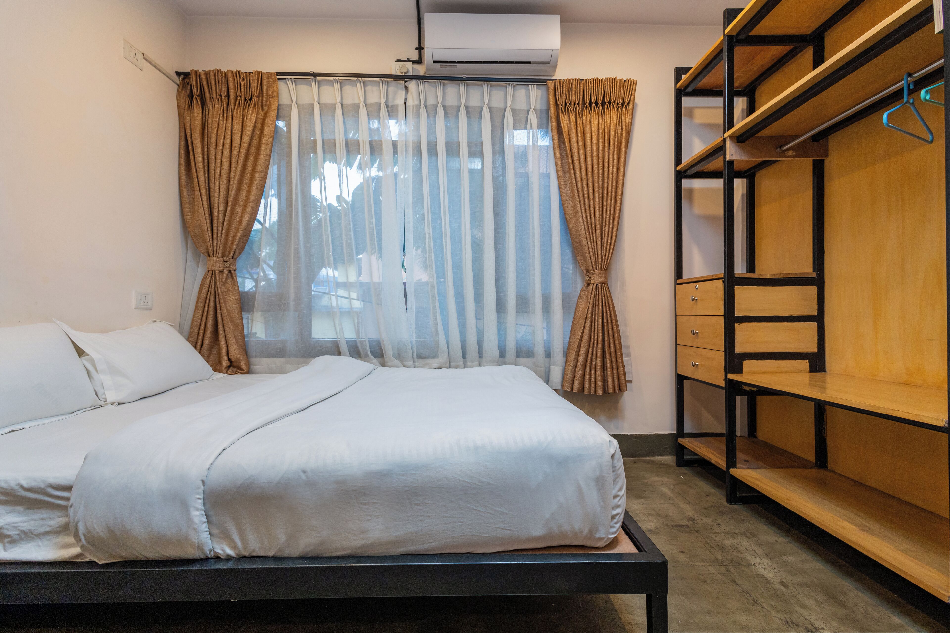A bedroom with a bed, curtains, an air conditioning unit, and a wooden shelf unit.