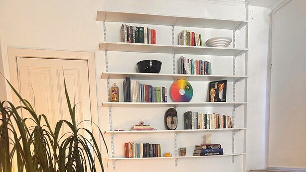 Een slaapkamer met een bed, een boekenplank, een geweven zitkussen en een plant.