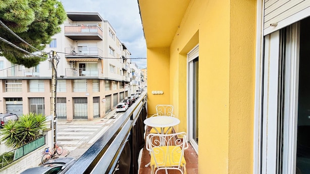 Un balcón con mesa y sillas, vista a una calle con coches estacionados y un edificio de varios pisos y balcones.