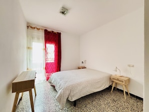 Un dormitorio con una cama, un escritorio, una lámpara y una ventana con cortinas rojas.