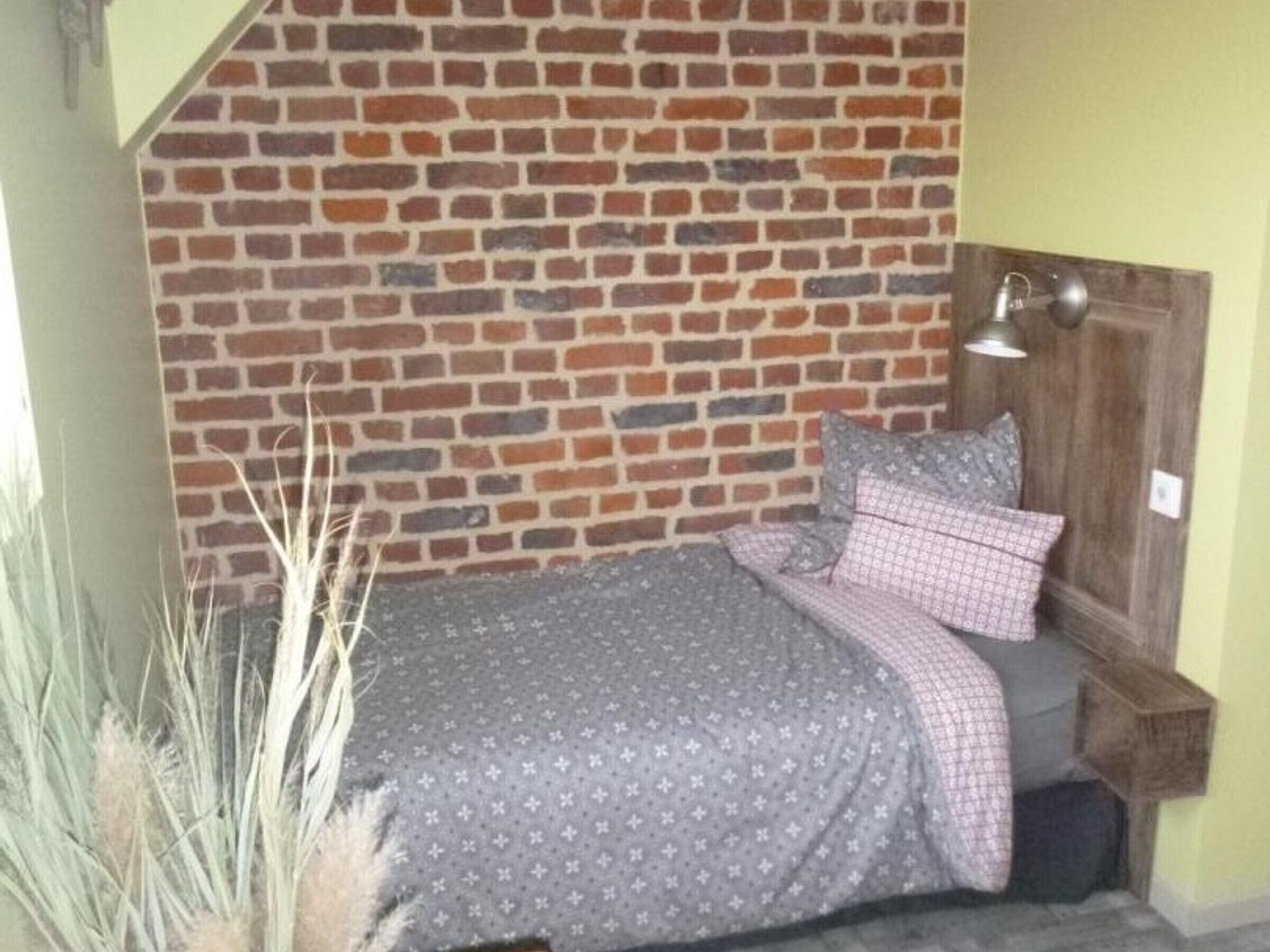 Une pièce avec un mur en briques, un lit recouvert d’une couvre-lit à motifs, des oreillers, une table de chevet et une lampe.