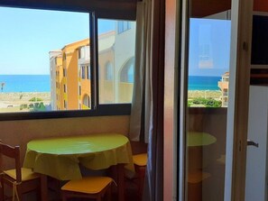 Ein Zimmer mit Tisch und Stühlen, ein Fenster mit Blick auf Strand und Meer sowie eine Tür zu einem weiteren Raum.