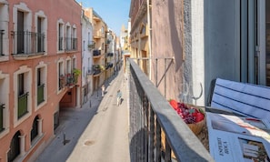 Eine schmale Straße mit Gebäuden an beiden Seiten, ein Fußgänger und eine Bank mit Obstschale.