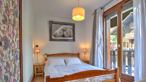 Een houten bed met witte beddengoed, twee bedlampjes, een lijst met een berenafbeelding, en uitzicht op een houten terras door een glazen deur.