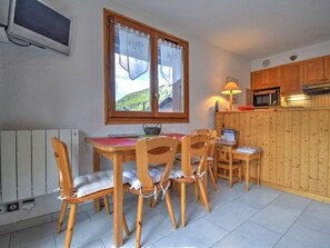 A dining area with a wooden table and chairs, a window with curtains, a lamp, and a kitchen area with wooden cabinets and appliances. - Charming apartment in Morzine - 4 guests (Morzine)