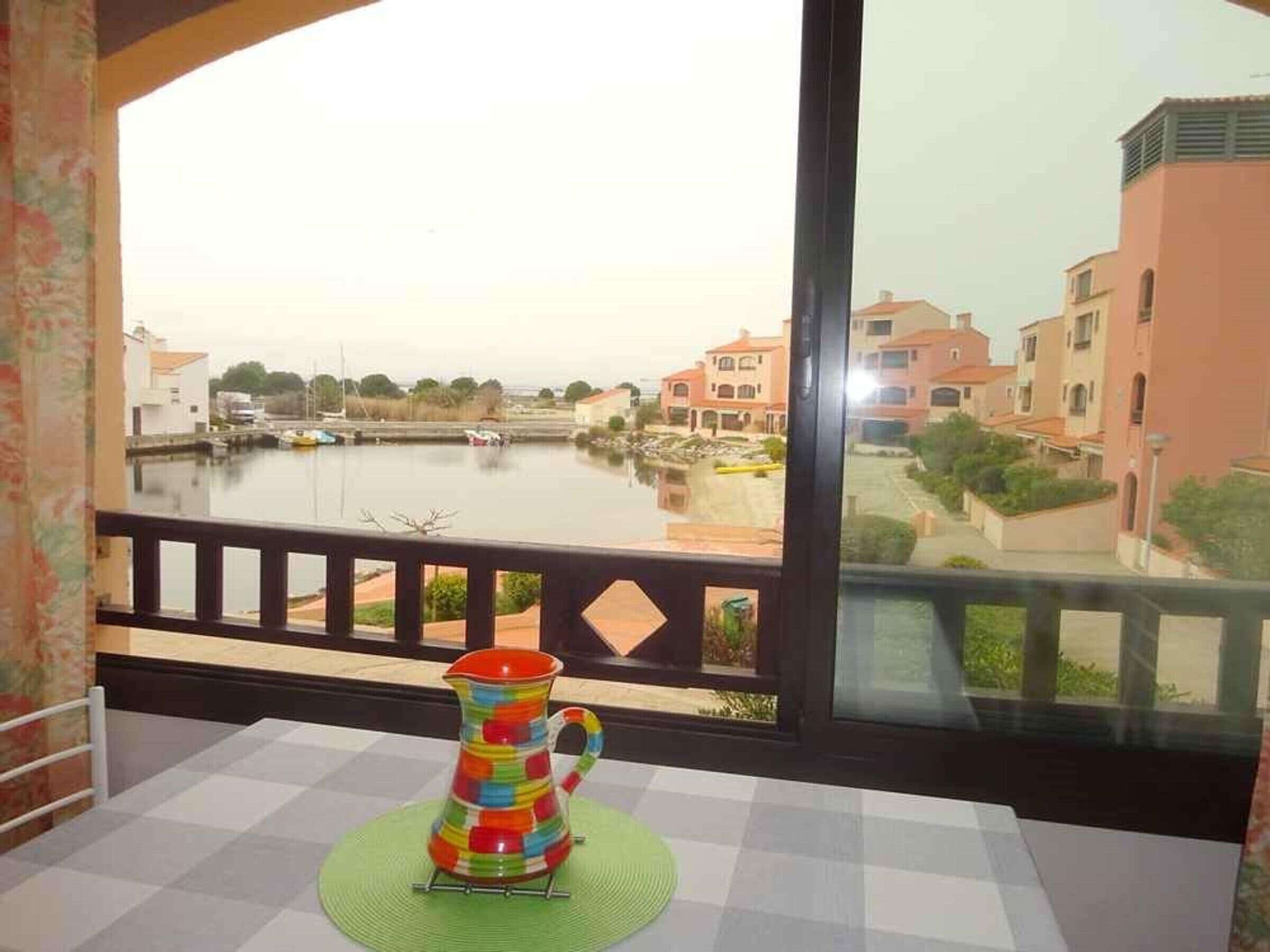 Una jarra colorida sobre una mesa, junto a una ventana con vistas a un canal y edificios.