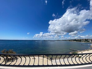 Ein Küstenblick mit klarem Himmel, ruhigem Meer und einer Promenade mit Geländer.