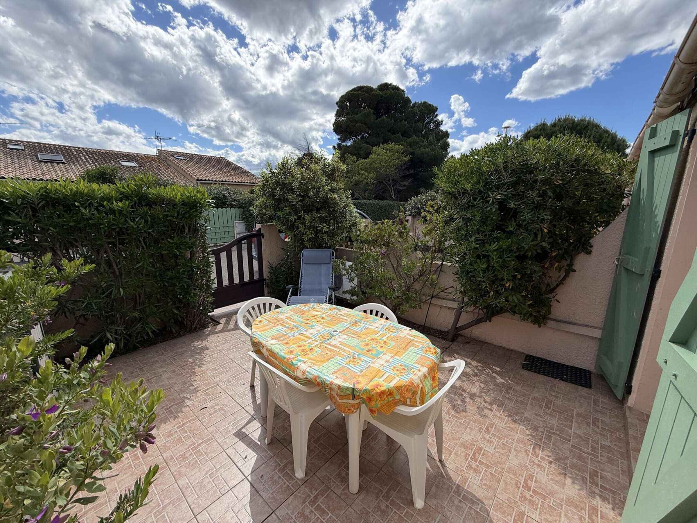 En terrasse med et bord og stole, omgivet af grøn vegetation og en bygning med et tag af fliser.
