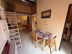 A compact living space with a loft area, a dining table with chairs, a red chair, a white door, and a kitchen area. - Villa with pool and AC - 4 guests (La Franqui)