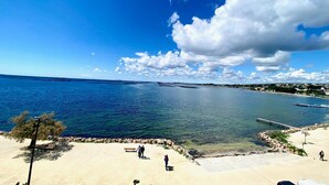 Vistes costaneres amb un cel serè, mar tranquil·la i una platja sorrenca amb persones i un moll.