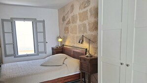 Un dormitori amb llit, una tauleta de nit, escriptori, làmpada i paret de pedra.