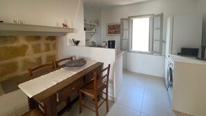 Una zona de menjador amb una taula i caires de fusta, una làmpada penjant, una paret de pedra i una cuina amb una màquina de rentar.