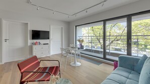 A modern living room with a sofa, a striped chair, a dining table with chairs, a flat-screen TV, and large windows with a view of trees and a balcony. - Żeromskiego 23 by Grand Apartments (Gdynia)