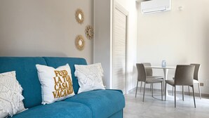 Een blauwe bank met decoratieve kussens, een eettafel met stoelen en wanddecoratie.
