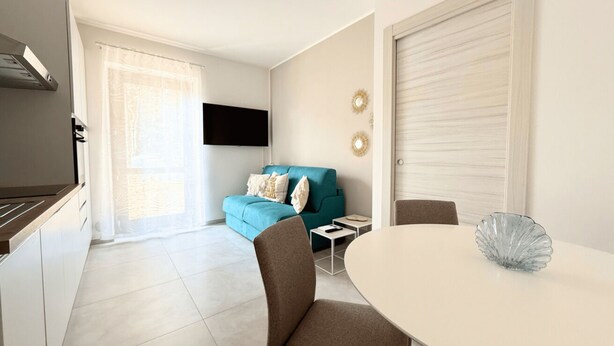 Een moderne keuken met een ingebouwde kookplaat, een eethoek met tafel en stoelen, een blauwe bank met kussens en een aan de muur bevestigde televisie.