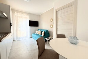 A modern kitchen with a built-in stove, a dining area with a table and chairs, a blue sofa with cushions, and a TV mounted on the wall. - S371 - Sirolo, apartment with garden and parking (Sirolo)