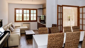 Ein Wohnzimmer mit Klavier, Sofa, Esstisch, Korbstühlen und einem Fernseher.