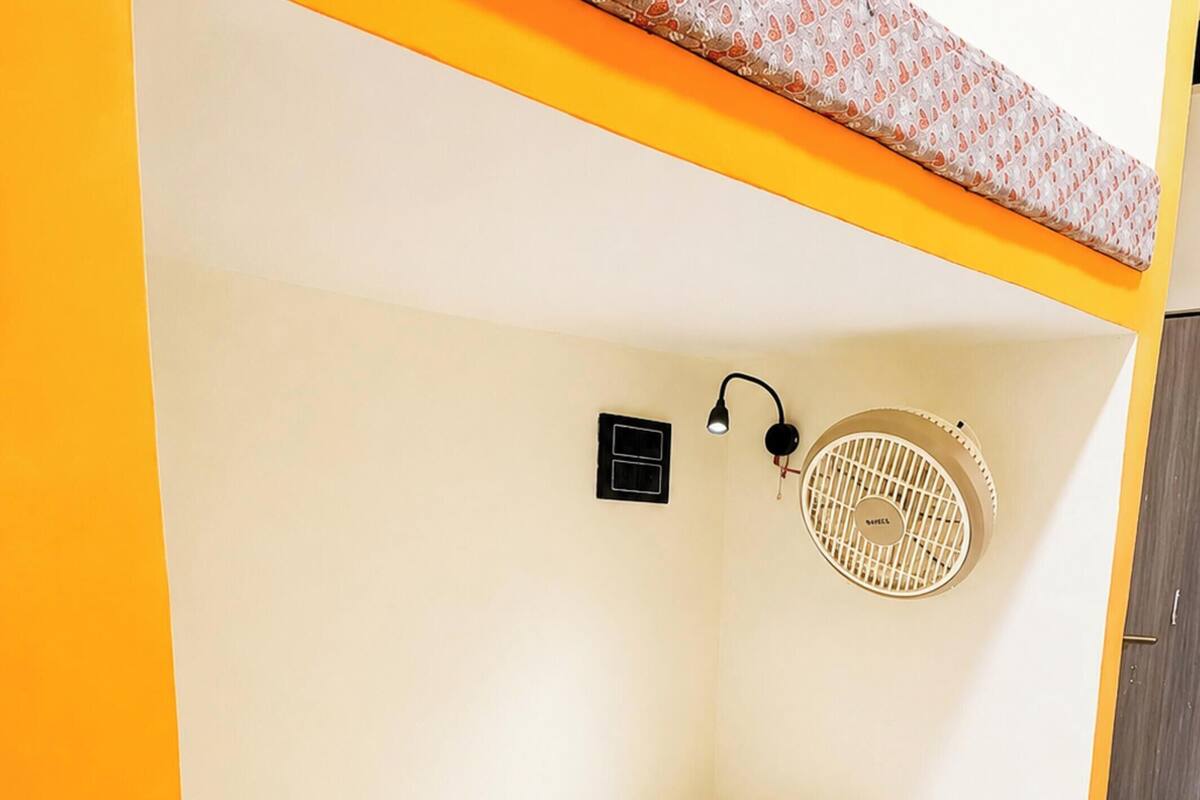 Una cama litera con colchón blanco, una manta estampada, un ventilador sujeto a la pared y un interruptor pequeño negro.