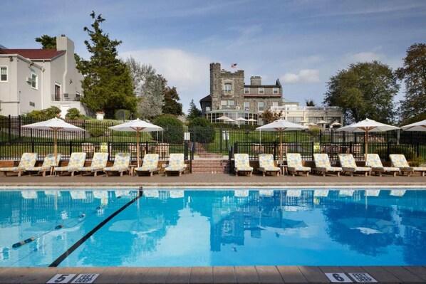 Una piscina con sillas de descanso y sombrillas, un edificio parecido a un castillo al fondo, y un cielo despejado.