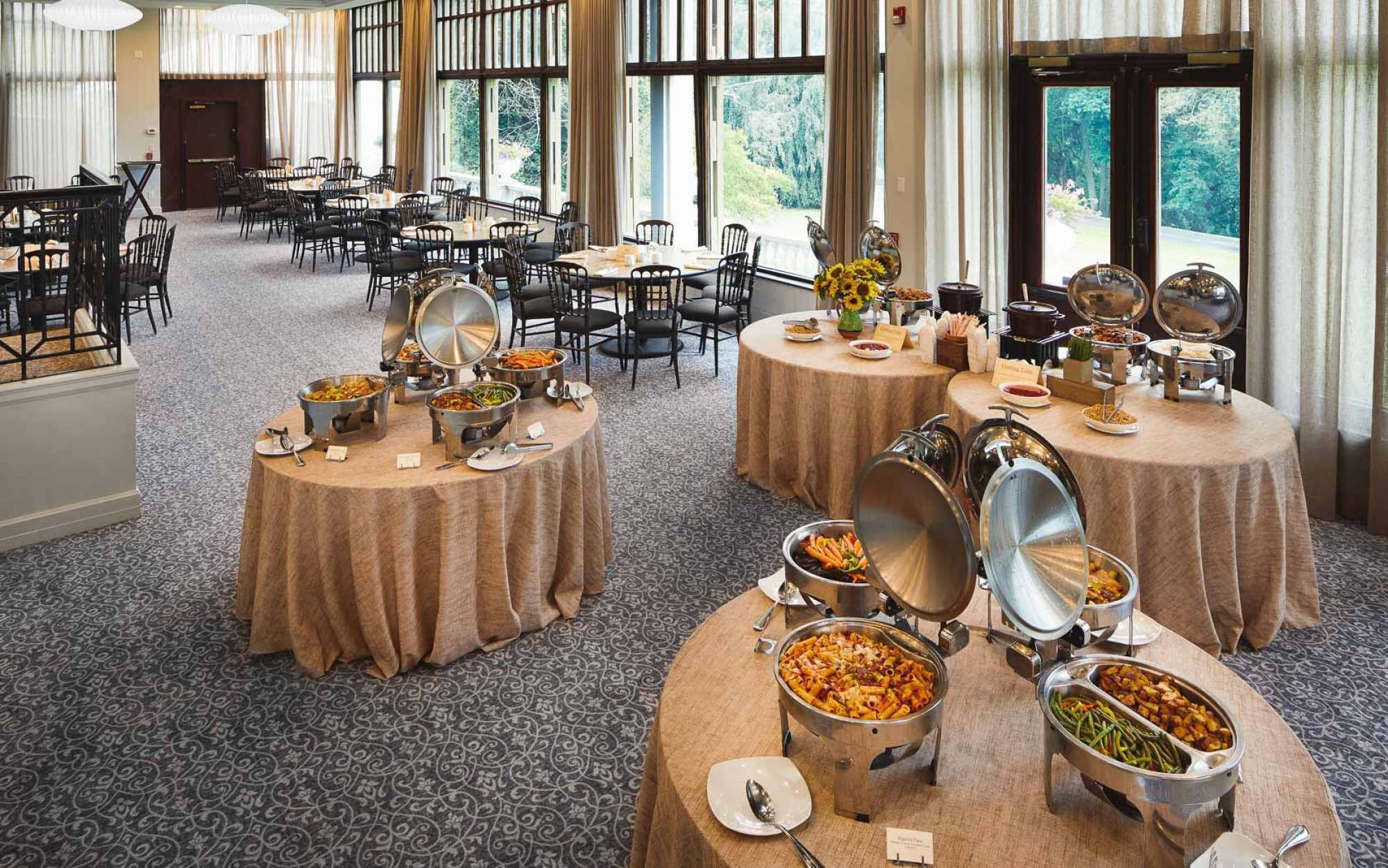 Un buffet composé de plusieurs tables rondes recouvertes de nappes, sur lesquelles sont disposés divers plats dans des récipients en métal.