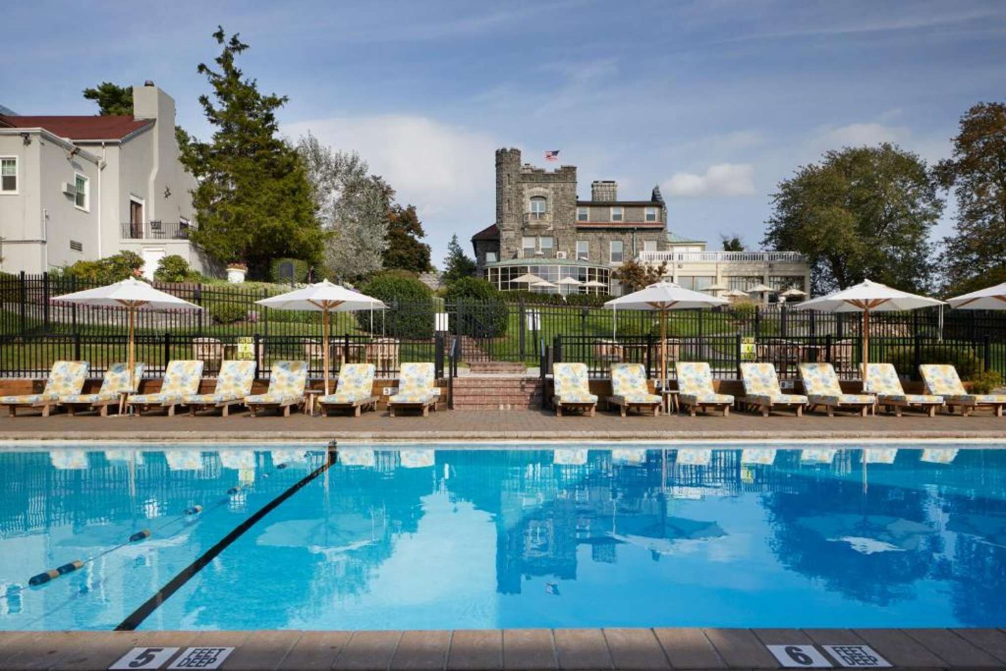 Une piscine avec des chaises longues et des parasols, un bâtiment qui ressemble à un château en arrière-plan, et un ciel dégagé.