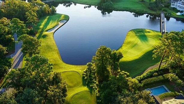 Eine Vogelperspektive auf einen Golfplatz mit Teich, umgeben von Bäumen und einer Wohngegend.