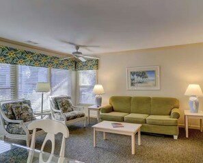 Ein Wohnzimmer mit einem grünen Sofa, weißen Sesseln, einem Glastisch und einem Deckenventilator.
