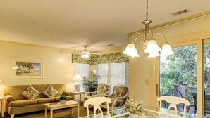 A living room with a glass dining table, white chairs, a sofa, and a ceiling fan.
