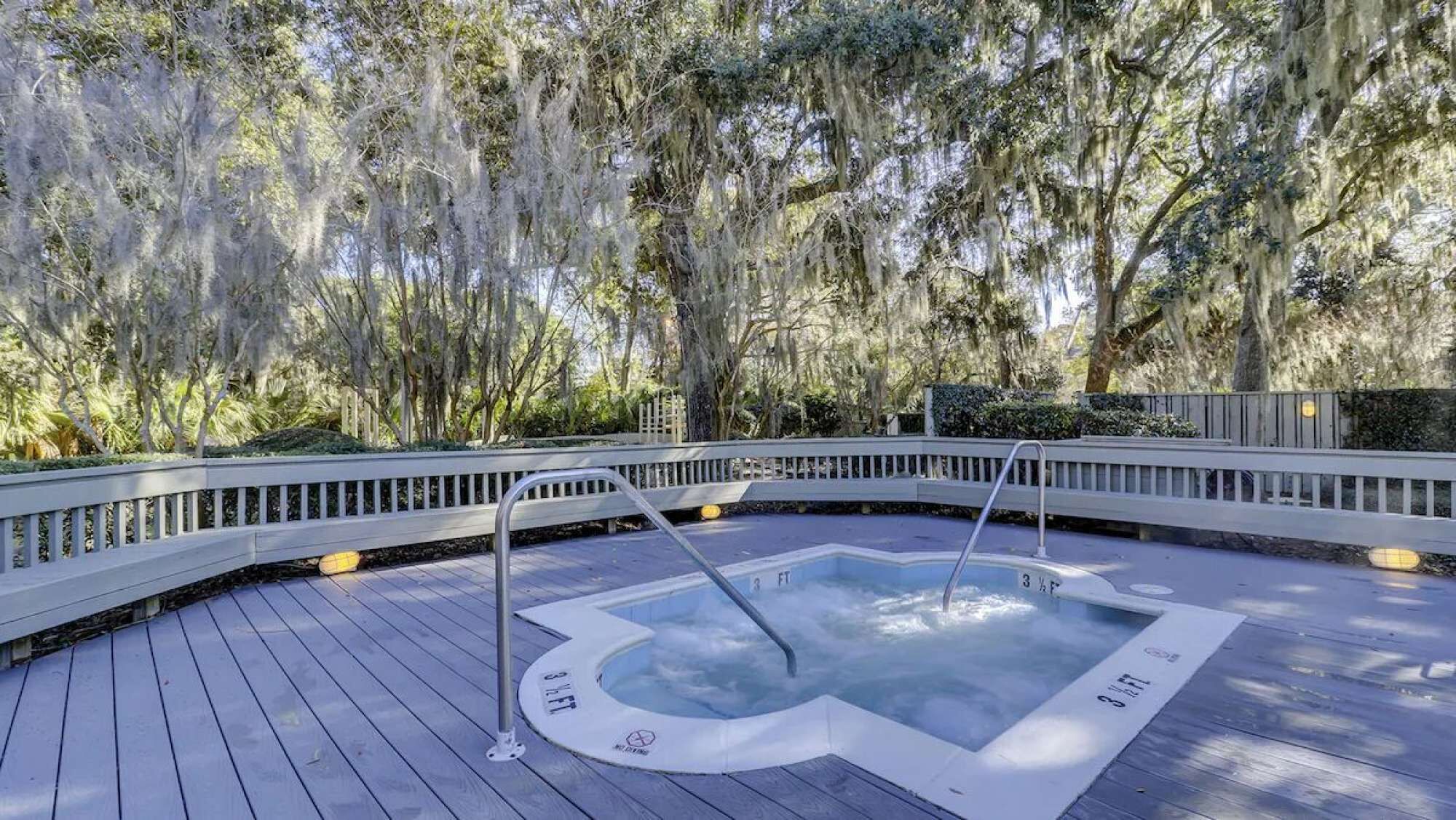 A hot tub on a wooden deck with a railing, surrounded by trees and greenery.