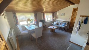 Ein gemütliches Zimmer mit Holzboden, einer Couch, einem Tisch mit Stühlen, einem Fernseher und einem Fenster mit Vorhängen.