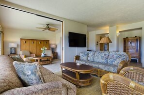 A living room with a sofa, chairs, a coffee table, a television, and a ceiling fan. - Prince Kuhio 108 - Spacious, A/C, close to beach (Koloa)