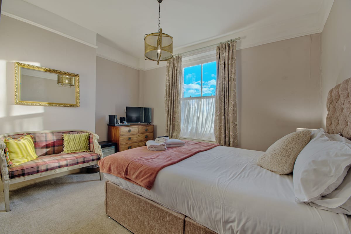 Quarto com cama, sofá, televisão, cómoda e janela com cortinas.