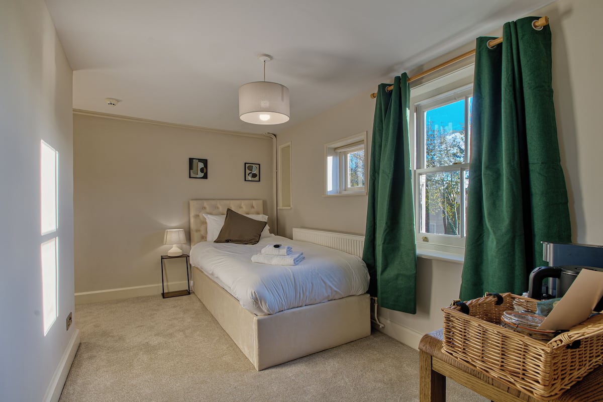 Quarto com cama, mesa de cabeceira, luminária, janela com cortinas e vista para árvores lá fora.