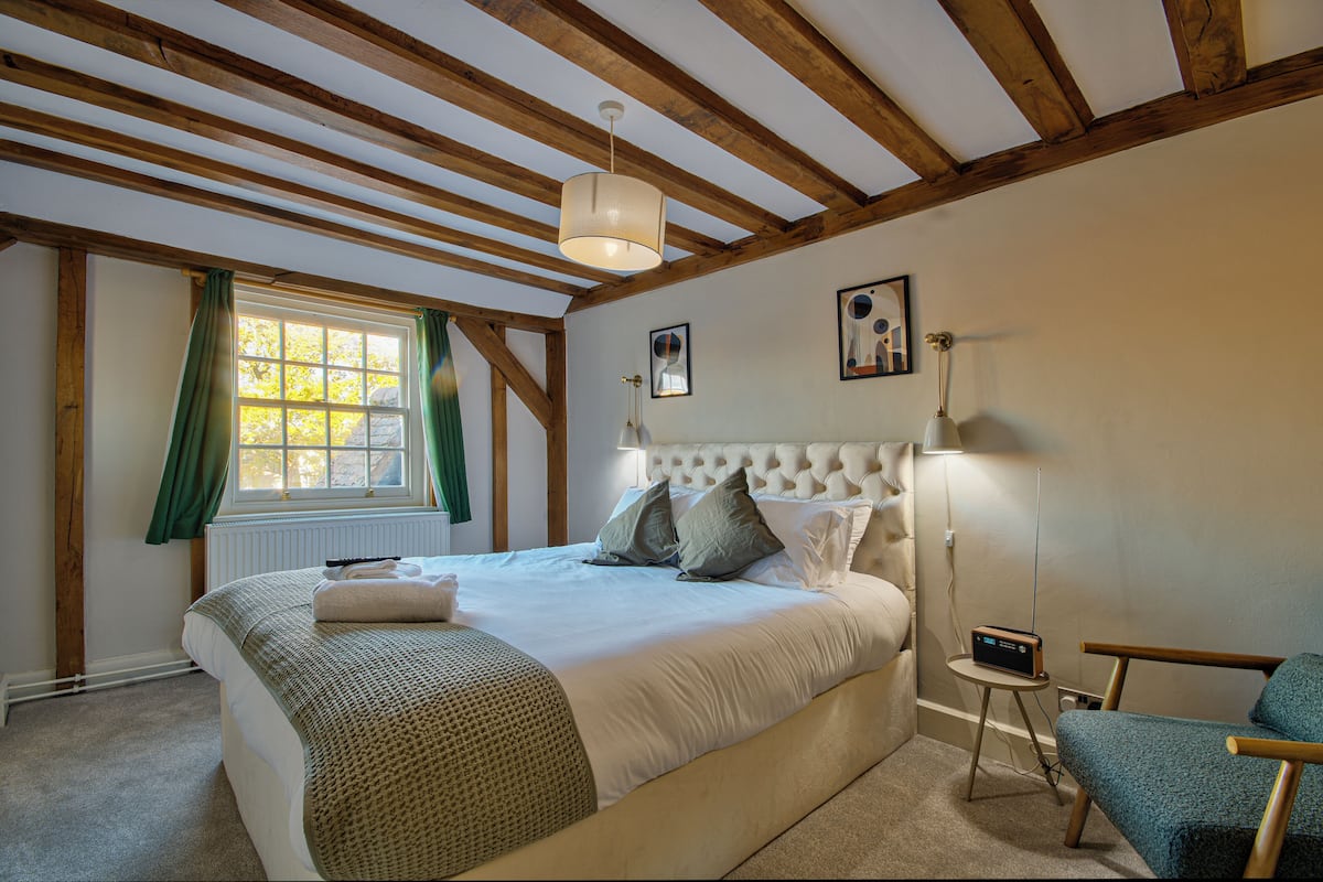 Quarto com cama grande, mesa de cabeceira, cadeira, janela com cortinas e teto com vigas de madeira.