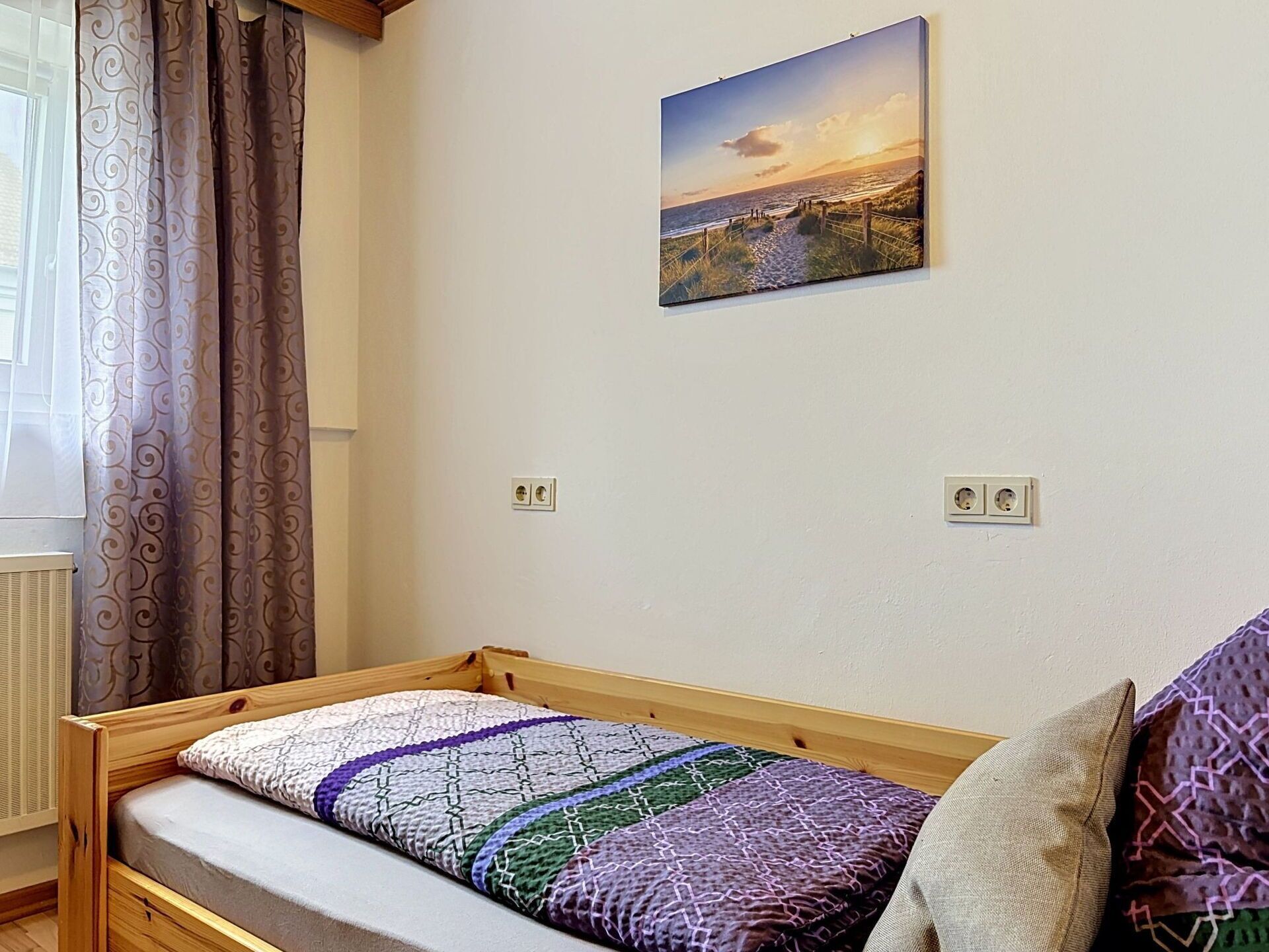 Ein Einzelbett mit einem gemusterten Bettdeckchen, ein Wandbild und ein Fenster mit Vorhängen.