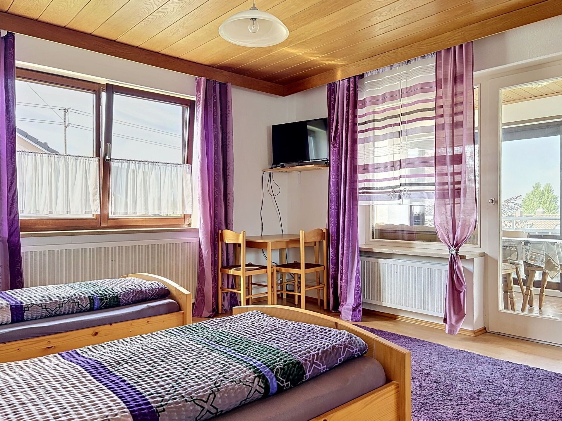 A room with two wooden beds, a desk, a chair, a TV, and a balcony with a view.