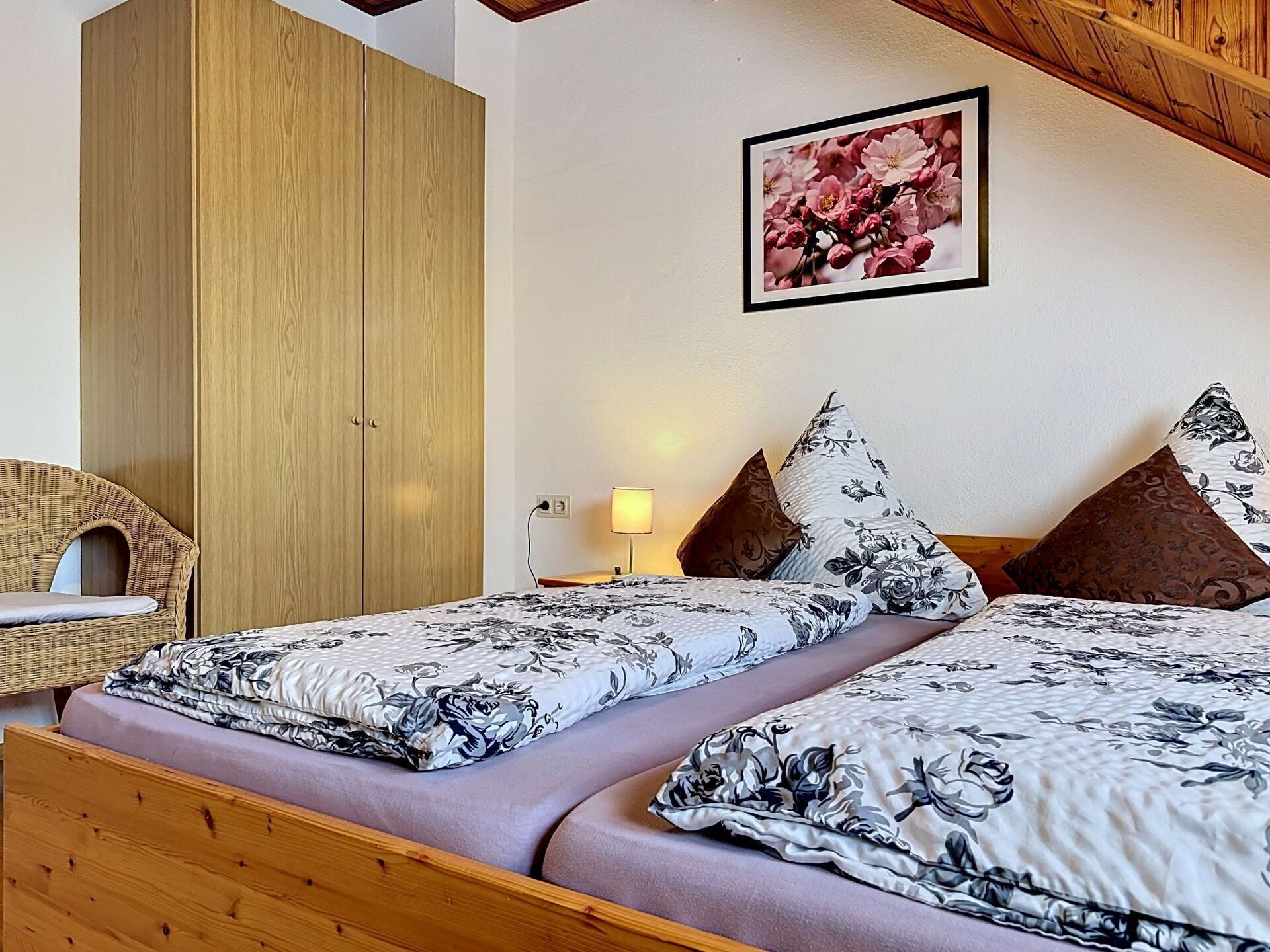 Ein Schlafzimmer mit zwei Betten, einem Holzschrank, einem Korbstuhl und einem Blumenbild an der Wand.