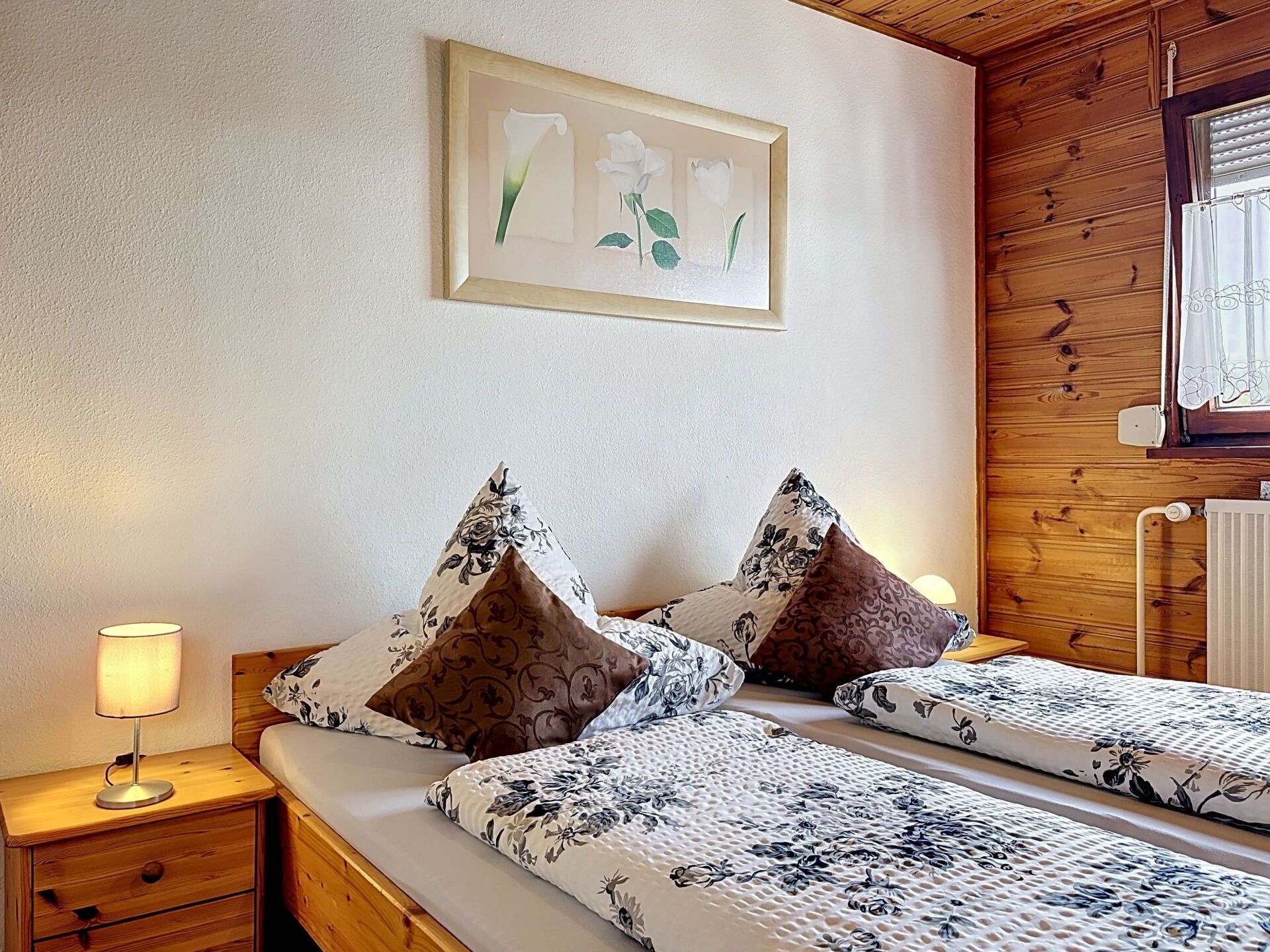 Een tweepersoonsbed met bloemenbeddengoed, een nachtkastje met een lamp, een kunstwerk met bloemen in een lijst aan de muur, en een houten wandpaneel.