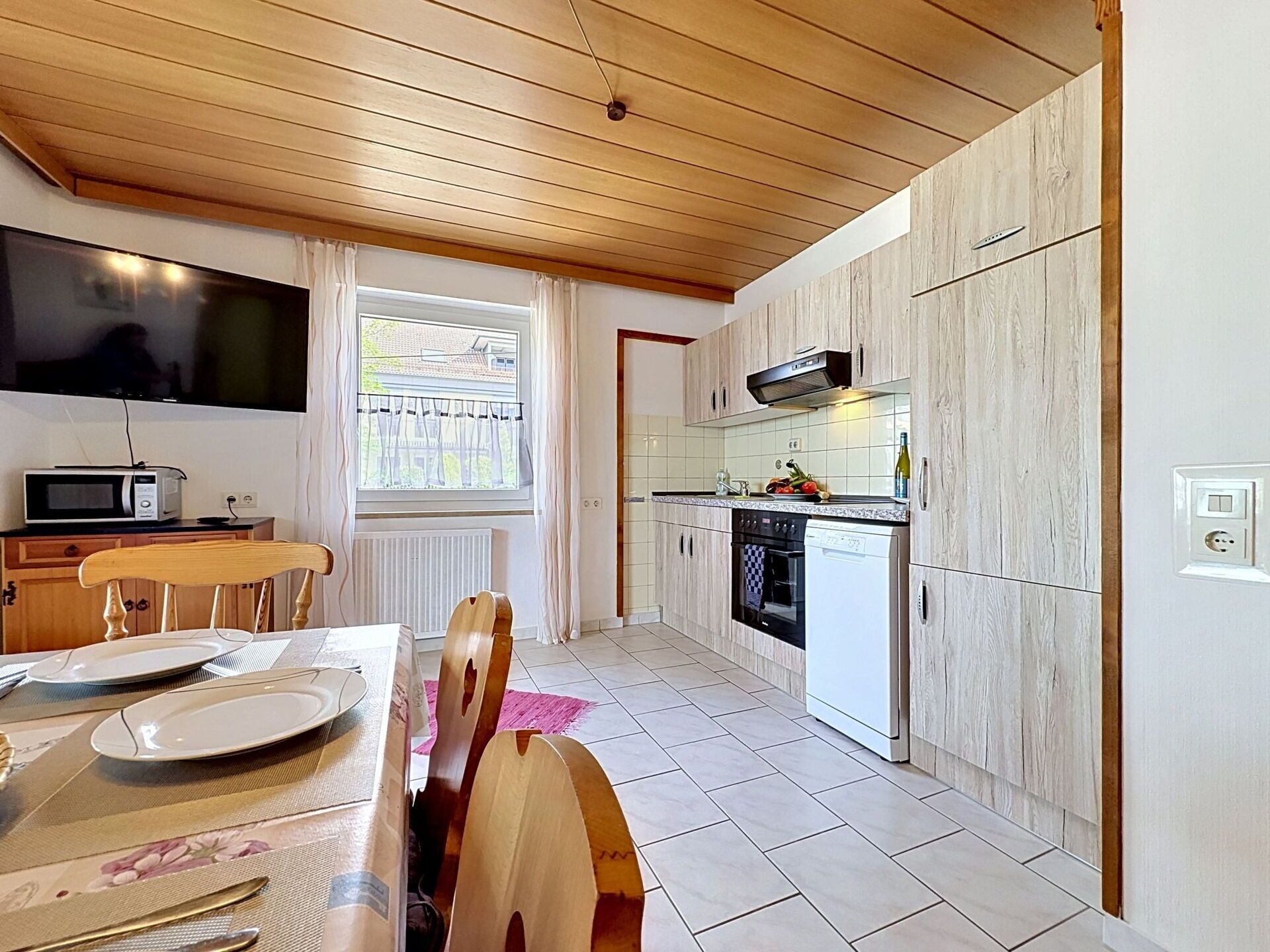 A modern kitchen with wooden ceiling, a dining area with a table set for two, and a window with curtains.