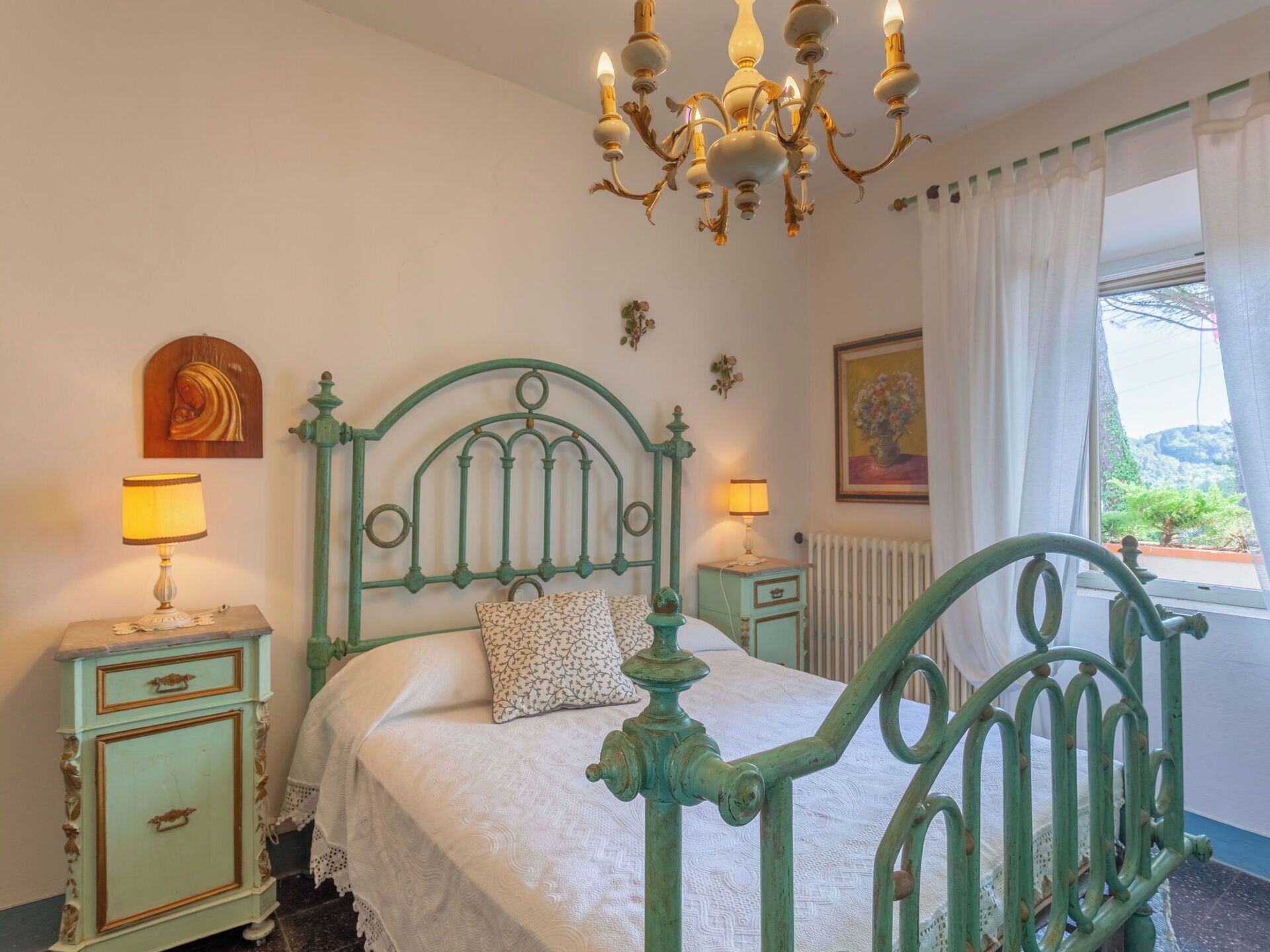 A bedroom with a green metal bed frame, two bedside tables with lamps, a chandelier, a radiator, and a window with curtains.