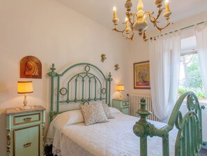 Ein Schlafzimmer mit einem grünem Metallbettgestell, zwei Nachttischlampen, einer Kristallleuchtreiter, einem Blumenbild und einem Heizkörper.